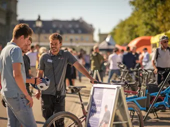 A man wearing a Bosch polo shirt stands in an urban setting with two men in front of an eBike, smiling at them. In the background, you can see pavilions and more eBikes.