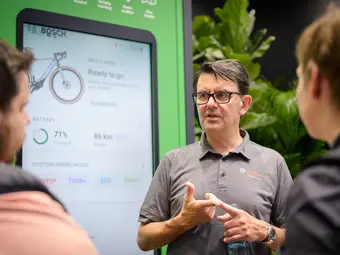 A man wearing a polo shirt with the Bosch logo stands in front of a poster featuring the layout of the Bosch eBike Flow app. He is explaining something to two people in front of the poster.