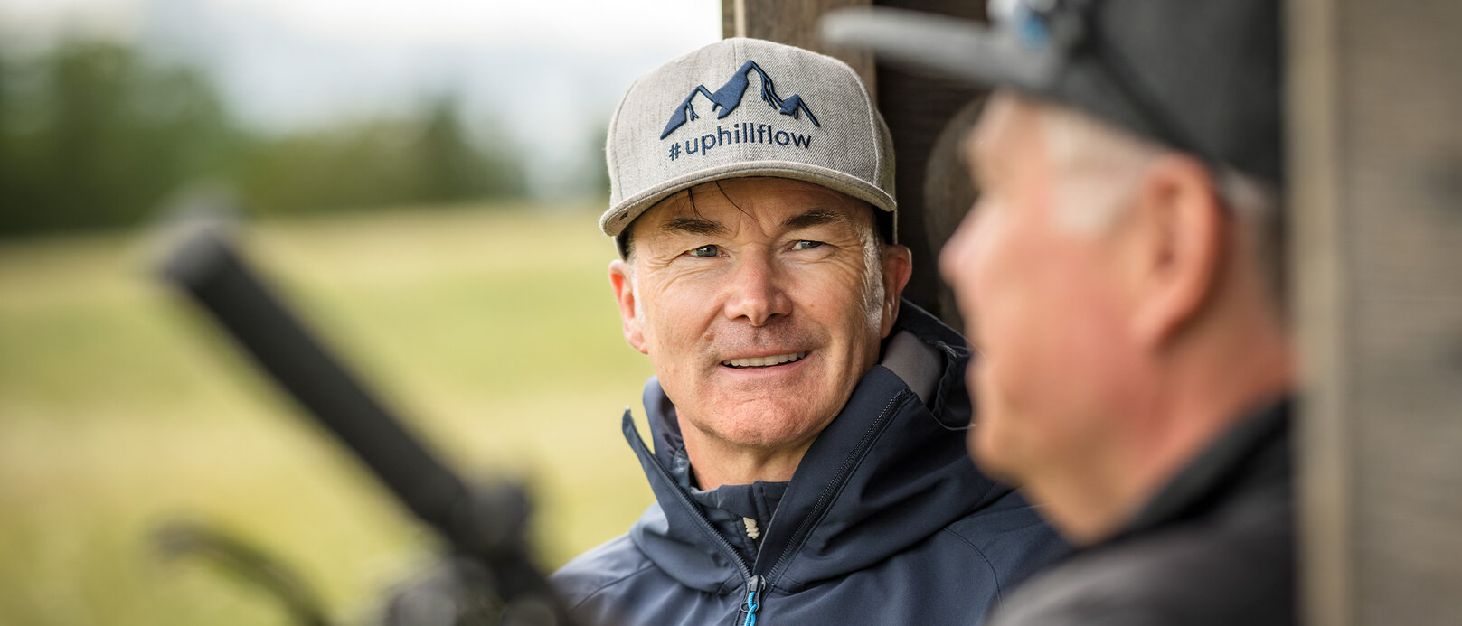Claus Fleischer porte une casquette sur laquelle est inscrit "#uphillflow" et une montagne est représentée. Il regarde une autre personne qui est floue à l'avant-plan.