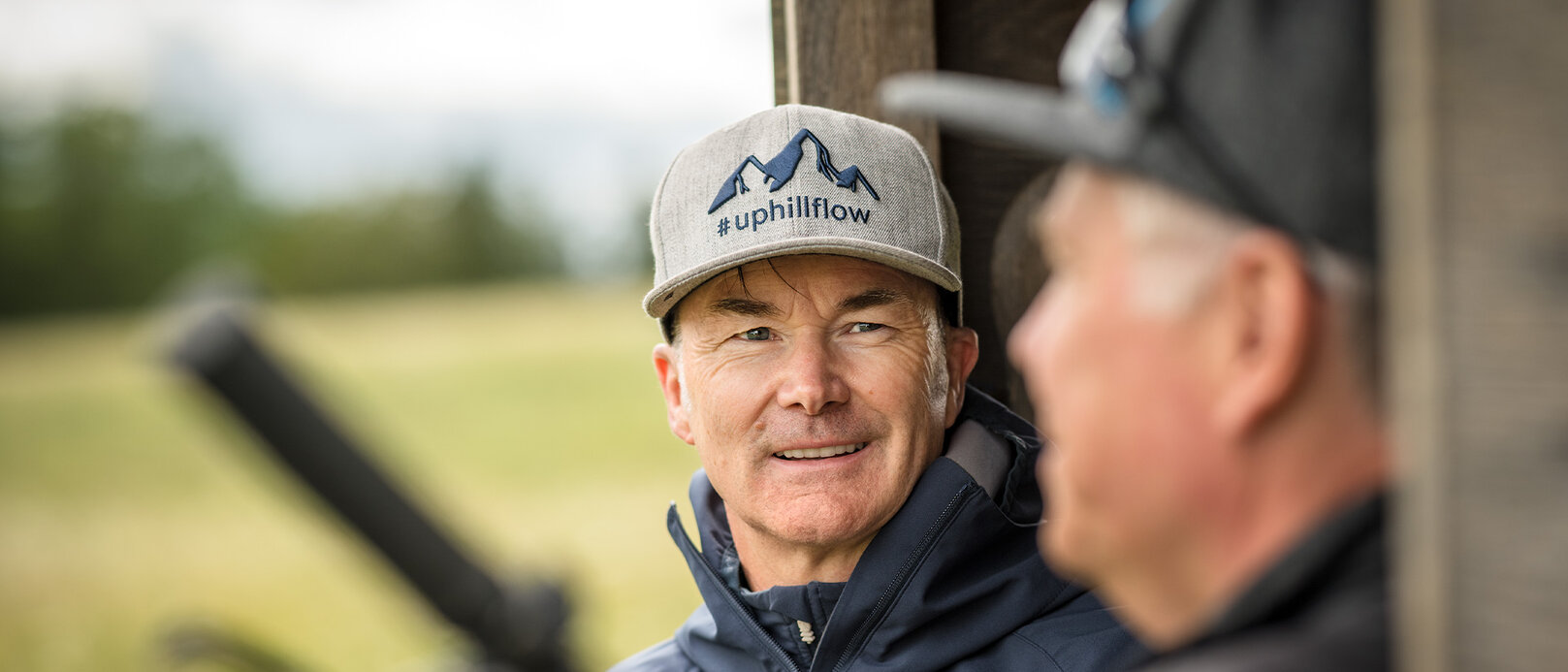 Claus Fleischer trägt eine Kappe, auf der "#uphillflow" steht und ein Berg abgebildet ist. Er schaut zu einer anderen Person, die im Vordergrund verschwommen zu sehen ist.