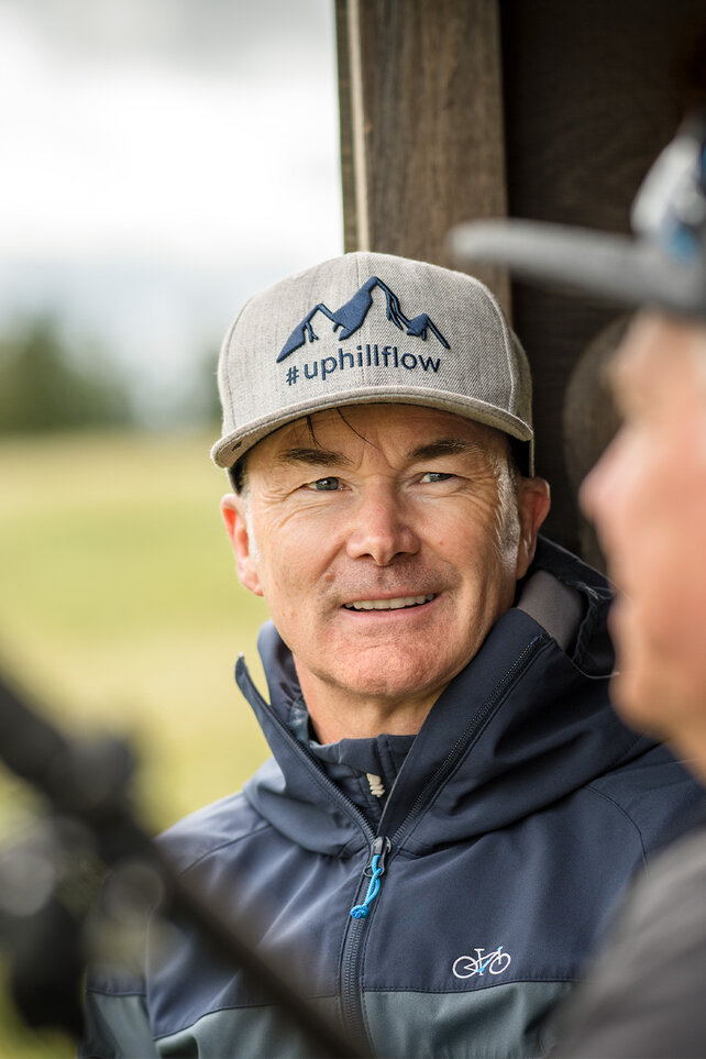 Claus Fleischer is wearing a cap with the inscription "#uphillflow" and a mountain depicted on it. He is looking at another person who appears blurred in the foreground.