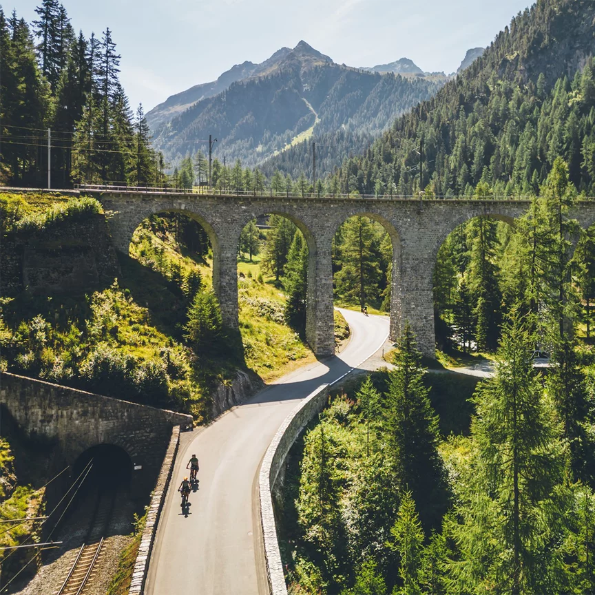 Deux conducteurs de VAE roulent le long d’une route qui passe sous un pont élevé. Autour d’eux, des montagnes recouvertes de forêt.