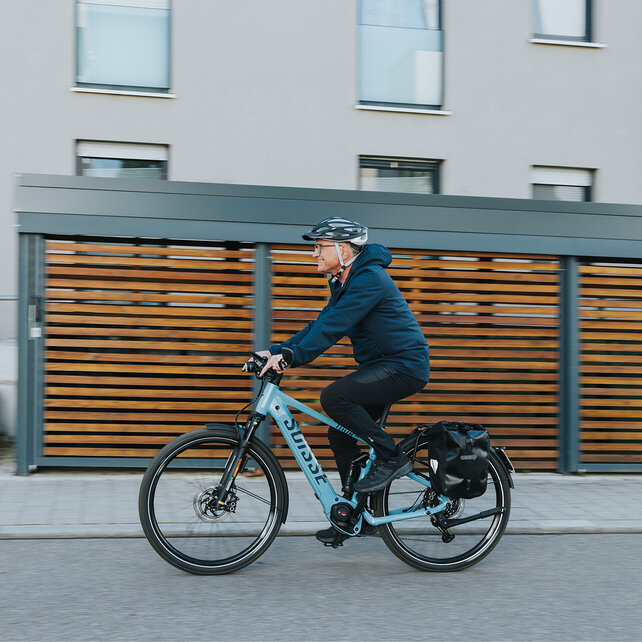 One person is riding a blue eBike and is wearing a black helmet and a dark blue jacket. In the background, you can see modern buildings and wood paneling.