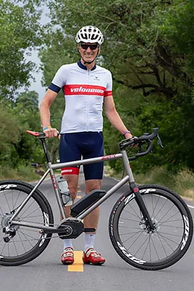 An eBike rider poses with his eBike across a road that leads through a forest.