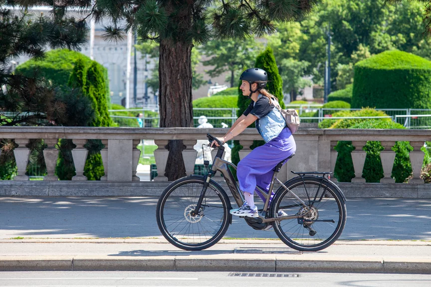 Eine Frau in einer weiten lila Hose fährt auf einem eBike in städtischer Umgebung einen Fahrradweg entlang. 