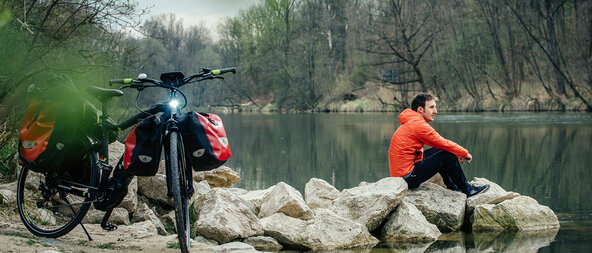 Ein eBiker sitzt an einem See, hinter ihm steht sein Trekking Pedelc mit Gepäcktaschen