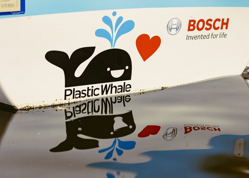 Close-up van een boot die op het water drijft. De boot heeft een blauw-witte print, een zwarte walvis met daaronder "PlasticWhale" en het rode Bosch-logo.