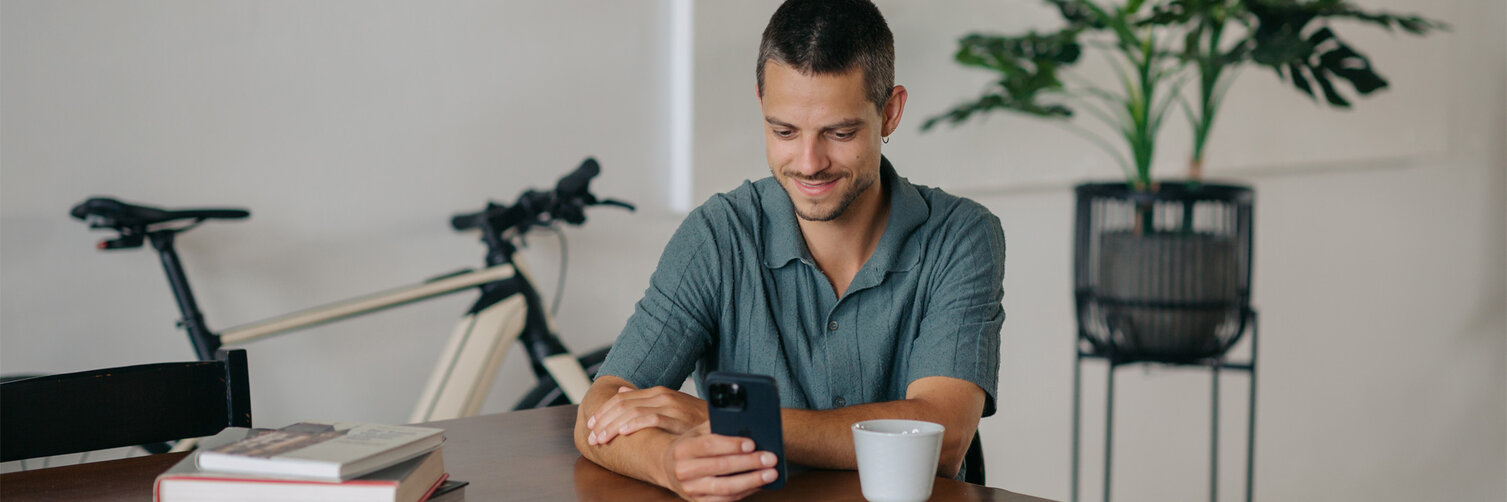 A man is sitting at a table, looking at his smartphone and smiling, with an eBike and a plant visible in the background.