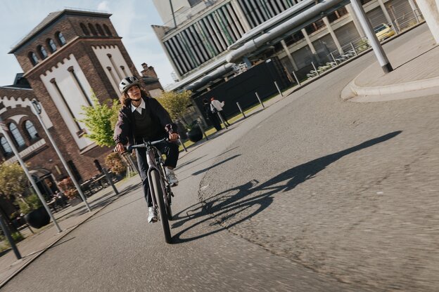 En kvinna med hjälm cyklar dynamiskt sin elcykel genom en kurva i ett urbant industriområde, förbi gammal och modern industriarkitektur.