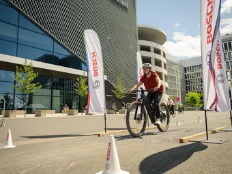 A woman rides an eBike along a street in front of a building complex. The route is marked with Bosch cones and Bosch flags.