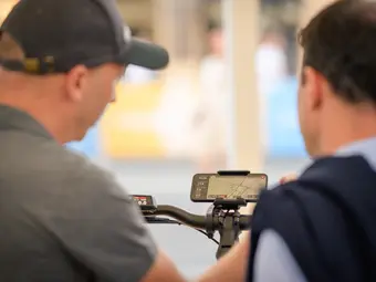 Two men look at a smartphone attached to the handlebars with a Smartphone Grip. The display shows active navigation in the eBike Flow app.