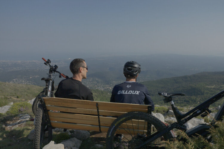 Vue de deux Vététiste assis sur un banc, de dos, contemplant un paysage de montagnes verdoyantes. Deux VTT sont garés à côté du banc.