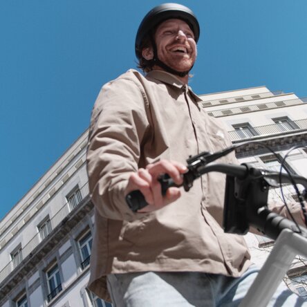 Desde la perspectiva de una rana: Un ciclista con casco sonríe con alegría al pasar junto a una fachada clásica y alta de edificio que resalta sobre un cielo azul brillante. La imagen transmite una sensación de libertad y felicidad en el entorno urbano.