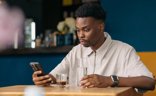 Mann sitzt in einem Restaurant und schaut auf sein Smartphone. Auf dem Tisch stehen Wasser und Kaffee.