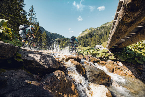 Dos ciclistas de eBike atraviesan un río en las montañas para llegar a la otra orilla.