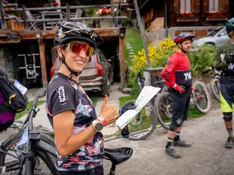 A woman in cycling gear is laughing, giving a thumbs up, and holding a map in her other hand. She is surrounded by other people with eBikes.