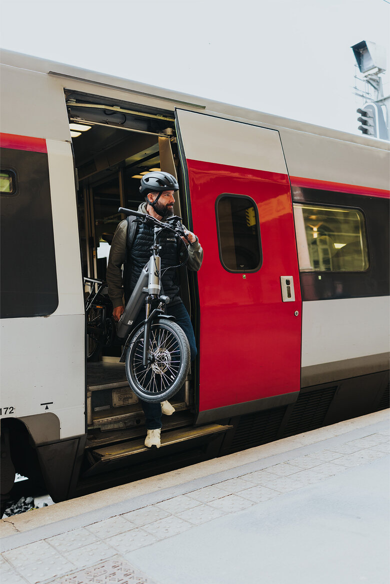 Eine Person steigt aus einem Zug und trägt ein schwarzes E-Bike. Die Person trägt einen schwarzen Helm und eine schwarze Jacke. Der Zug hat eine rot-graue Lackierung und steht am Bahnsteig.