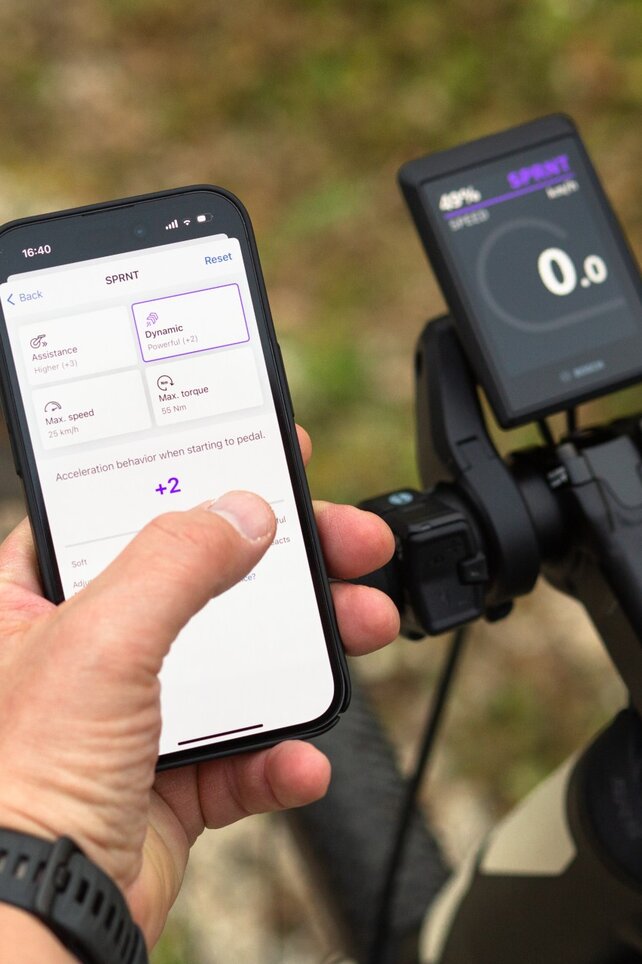 A person holds a smartphone displaying the ebike flow app with settings for support, speed, maximum torque, and acceleration behavior. The smartphone is held next to a bicycle handlebar, with a slightly blurred background showing an outdoor environment.