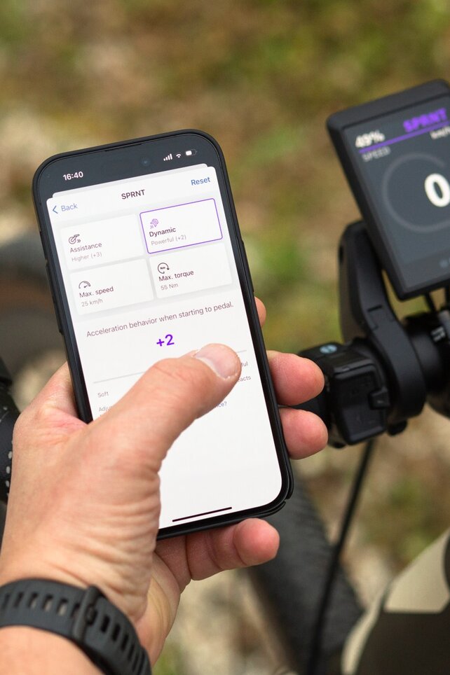 A person holds a smartphone displaying the ebike flow app with settings for support, speed, maximum torque, and acceleration behavior. The smartphone is held next to a bicycle handlebar, with a slightly blurred background showing an outdoor environment.