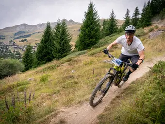 A cyclist rides along a narrow path, leaning slightly into the curve. In the background, a valley and trees are visible.