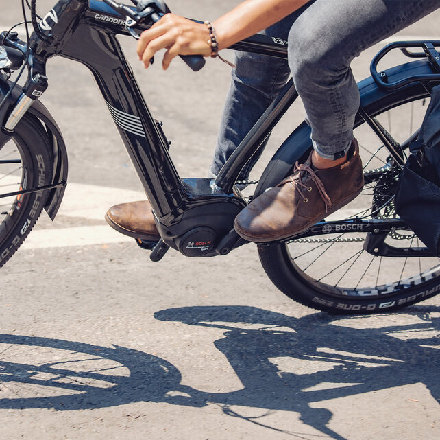 A close-up of an eBike in motion, ridden by a person wearing brown shoes and gray jeans. The eBike is equipped with a Bosch Performance Line Sport motor. The background shows a paved road with markings.
