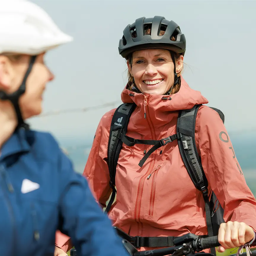 Two people are wearing bicycle helmets and outdoor clothing. One is dressed in a blue jacket with a white helmet, while the other wears an orange jacket with a black helmet. Both have backpacks and are in a rural environment.