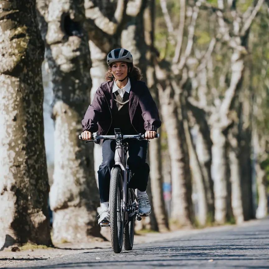 Una donna sorridente con un casco da bicicletta pedala sul suo eBike, equipaggiata con un sistema di eBike Bosch, lungo un viale soleggiato fiancheggiato dagli alberi.