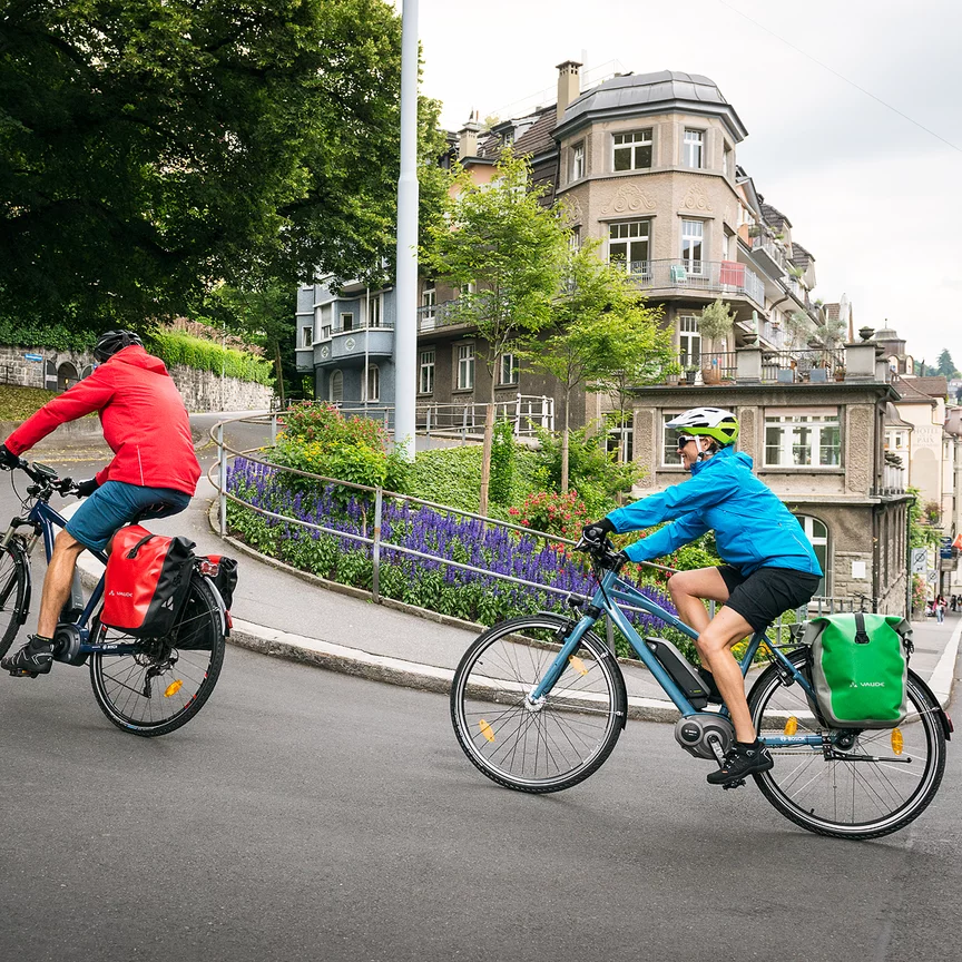 Two eBike riders are cycling uphill on eBikes along a steep street in a city.