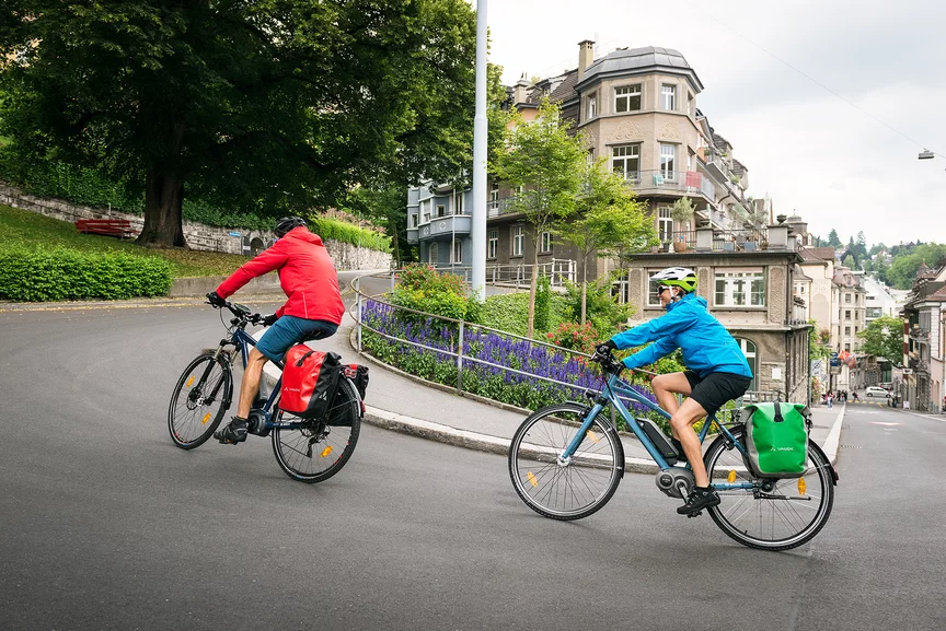 Zwei eBike Fahrer fahren auf eBikes eine steile Straße in einer Stadt hinauf.