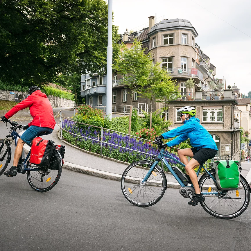 Zwei eBike Fahrer fahren auf eBikes eine steile Straße in einer Stadt hinauf.