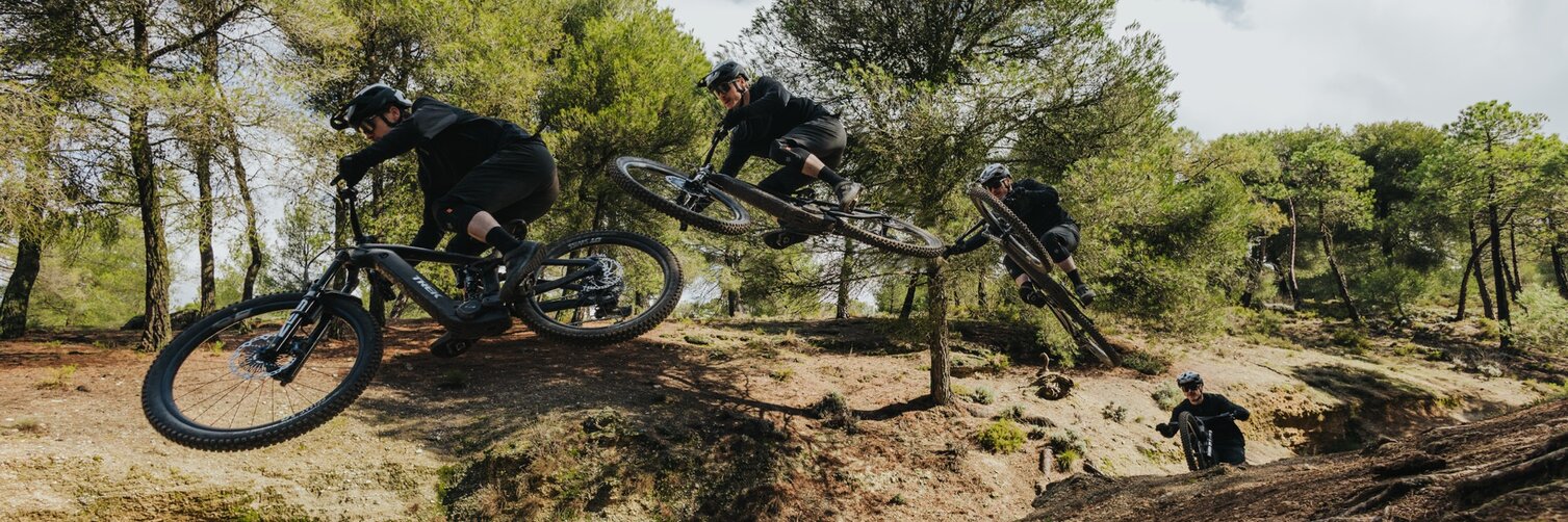 Sequenzaufnahme eines eMountainbikers, der einen Sprung auf einem staubigen Wald-Trail vollführt. Die Aufnahme zeigt den Fahrer in drei verschiedenen Phasen des Sprungs – vom Absprung über die Flugphase bis zur Landung.