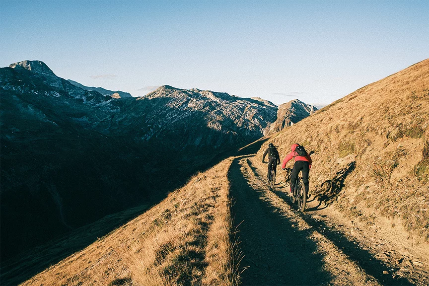 Deux conducteurs de VAE gravissent une montagne sur un chemin herbeux, avec d'autres montagnes visibles à l'arrière-plan.