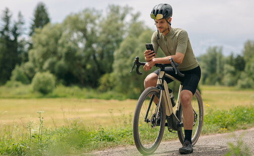 Mann macht eine Pause auf seinem eBike, schaut in sein Smartphone. Im Hintergrund sind Wiesen und Wald zu sehen.