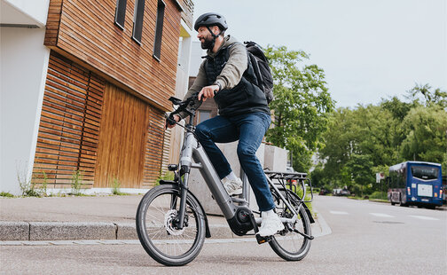 En person sykler på en grå elsykkel og har på seg en svart hjelm og en svart ryggsekk. Personen er på vei langs en gate ved siden av en moderne bygning med trepanel. I bakgrunnen ser du trær og en blå buss.