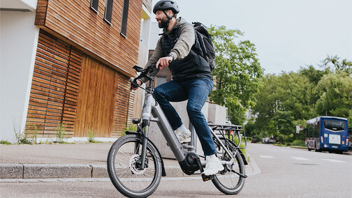 A person is riding a gray e-bike, wearing a black helmet and a black backpack. They are on a street next to a modern building with wood paneling. In the background, you can see trees and a blue bus.