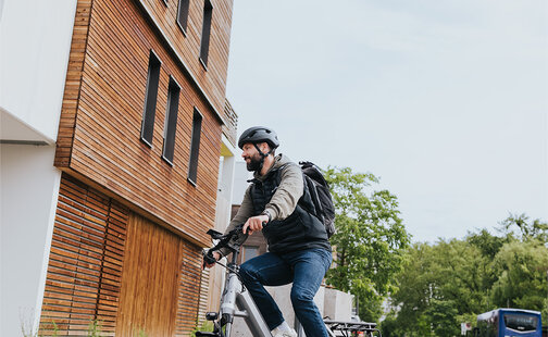 Eine Person fährt ein graues E-Bike und trägt einen schwarzen Helm sowie einen schwarzen Rucksack. Die Person ist auf einer Straße unterwegs, neben einem modernen Gebäude mit Holzverkleidung. Im Hintergrund sind Bäume und ein blauer Bus zu sehen.