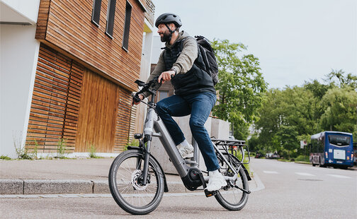 Une personne circule sur un vélo électrique gris et porte un casque noir ainsi qu’un sac à dos noir. Elle se trouve dans une rue, à côté d’un bâtiment moderne avec des boiseries. En arrière-plan, on distingue des arbres et un bus bleu.