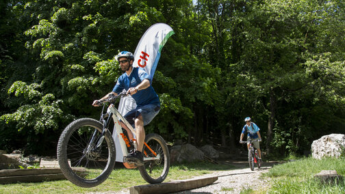 Un homme roule sur une poutre en bois sur un VTT à assistance électrique, tandis qu’une deuxième personne s’approche de l’obstacle derrière lui. À côté de l’obstacle se trouve un drapeau Bosch ; en arrière-plan, la forêt.