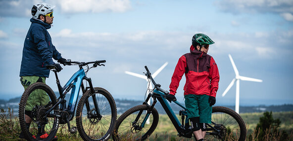 2 men are taking a rest while leaning on their eBikes, in front of wind power generators