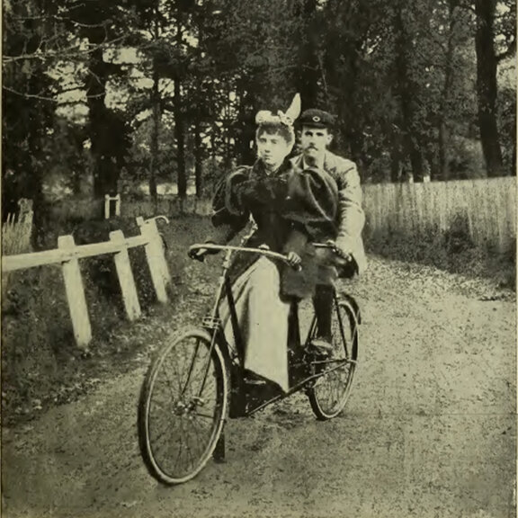 Foto in bianco e nero di un uomo e una donna che pedalano su una bicicletta tandem attraverso un bosco.