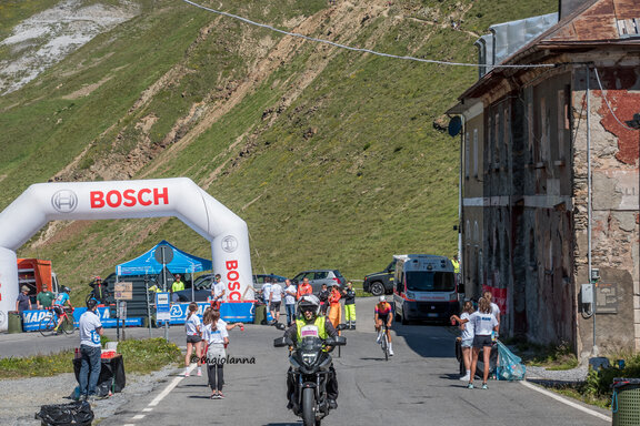 Rettifilo d’arrivo di una gara ciclistica: arco d’arrivo Bosch gonfiabile attorno al quale si trovano organizzatori, personale di sicurezza e altri volontari. Un motociclista con giubbotto ad alta visibilità si avvicina alla telecamera.