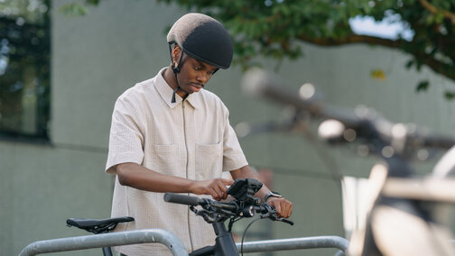 A man stands at a bicycle rack, gripping the handlebars of his eBike and tapping on the smartphone mounted in the smartphone grip holder on the eBike.