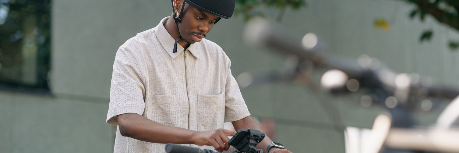 A man stands at a bicycle rack, gripping the handlebars of his eBike and tapping on the smartphone mounted in the smartphone grip holder on the eBike.