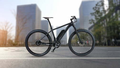 Černé eBike / elektrokolo stojí na ulici, v pozadí jsou vidět budovy a stromy.
