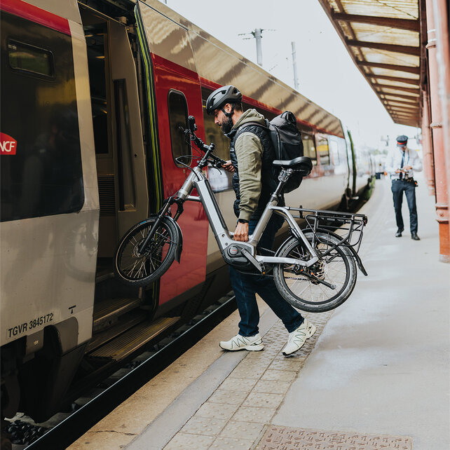 Un hombre barbudo con casco de bicicleta y mochila sube a un tren llevando una eBike plegable plateada.