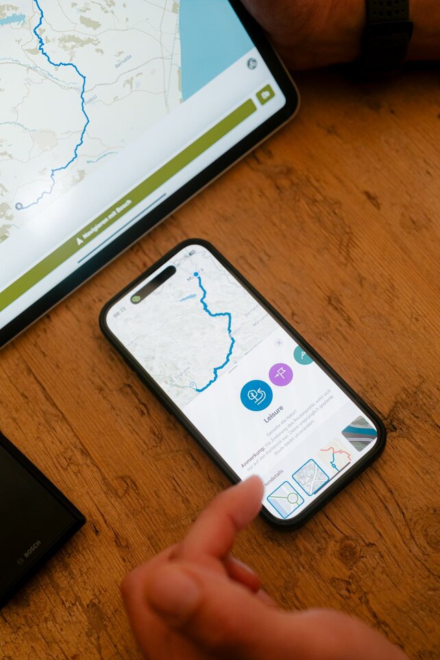 A smartphone with the eBike Flow app lies on a table. It displays a map with a marked route. Next to the smartphone is a tablet that also shows a map with a route.