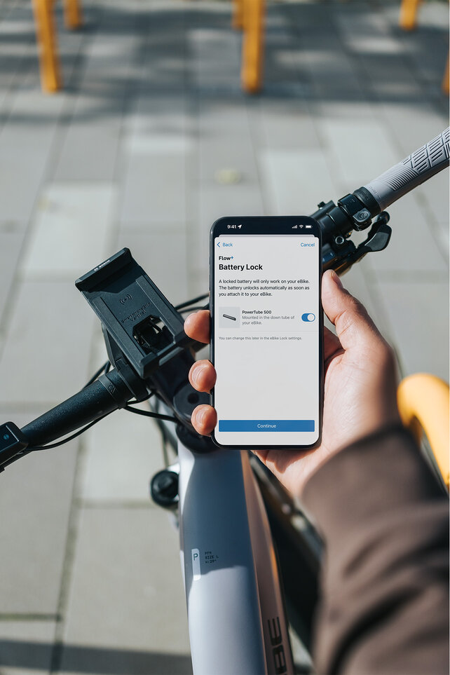 Focus on a smartphone being held in the hand. The screen displays the FlowApp with the BatteryLock function activated. In the background, you see an eBike handlebar with SmartphoneGrip Vertical.