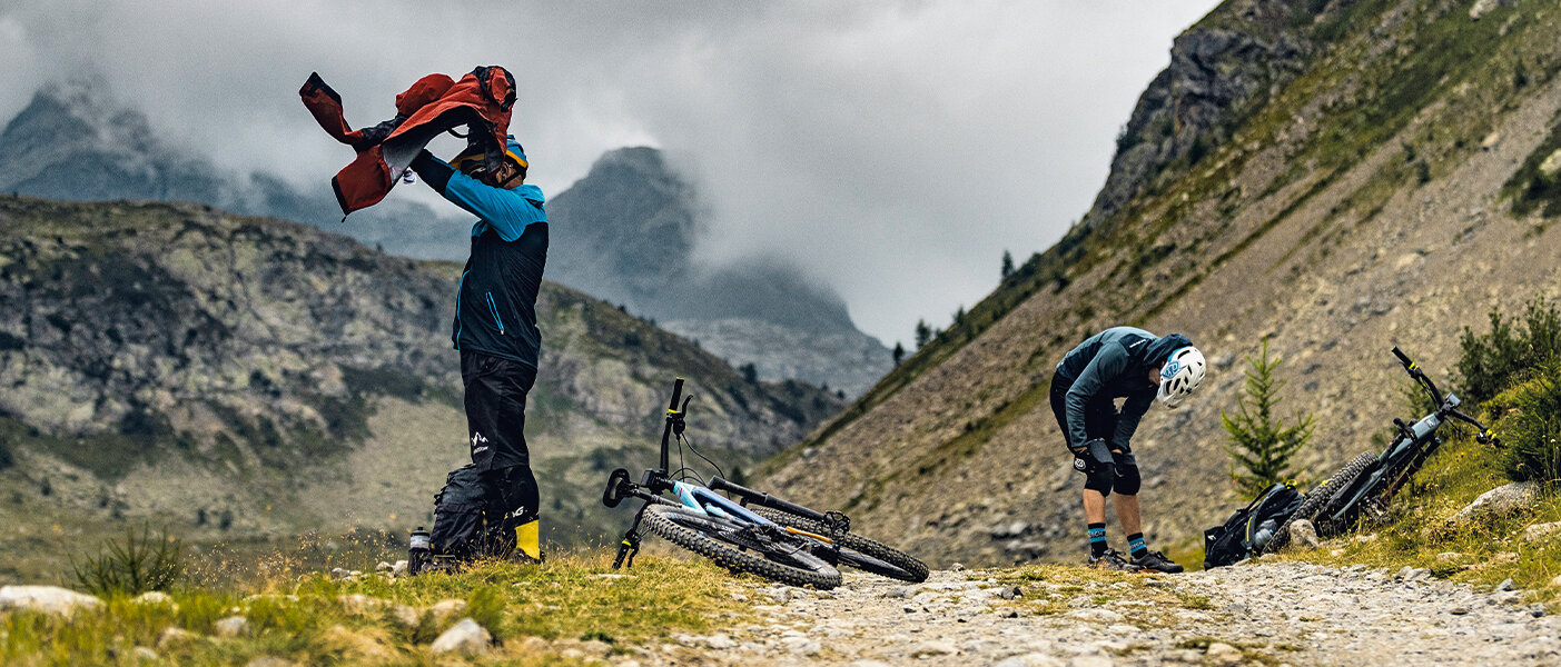 Dos ciclistas de eBike se están vistiendo de nuevo con su ropa de ciclismo en una ruta de montaña, mientras sus eBikes están apoyadas en el borde del camino.