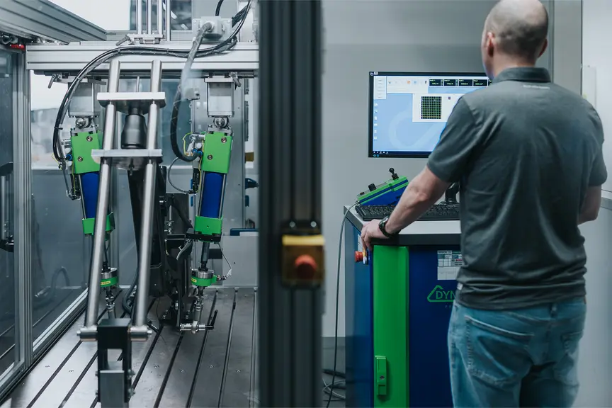 Eine Person bedient eine Maschine in einer industriellen Umgebung. Die Maschine hat grüne und blaue Komponenten, und die Person steht vor einem Computerbildschirm, der technische Daten anzeigt.