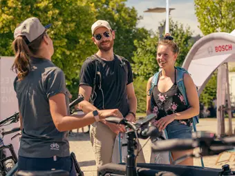 A Bosch expert is speaking with two smiling individuals, surrounded by bicycles, with a Bosch tent and trees visible in the background.